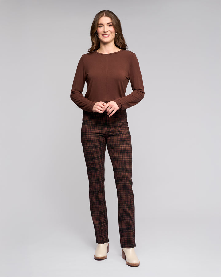 A woman with long brown hair stands smiling against a gray background, wearing Vassalli's Bay Pull On - Cherry slim plaid trousers with an elasticated waistband, a brown top, and cream heeled boots. Her hands are gently clasped in front.