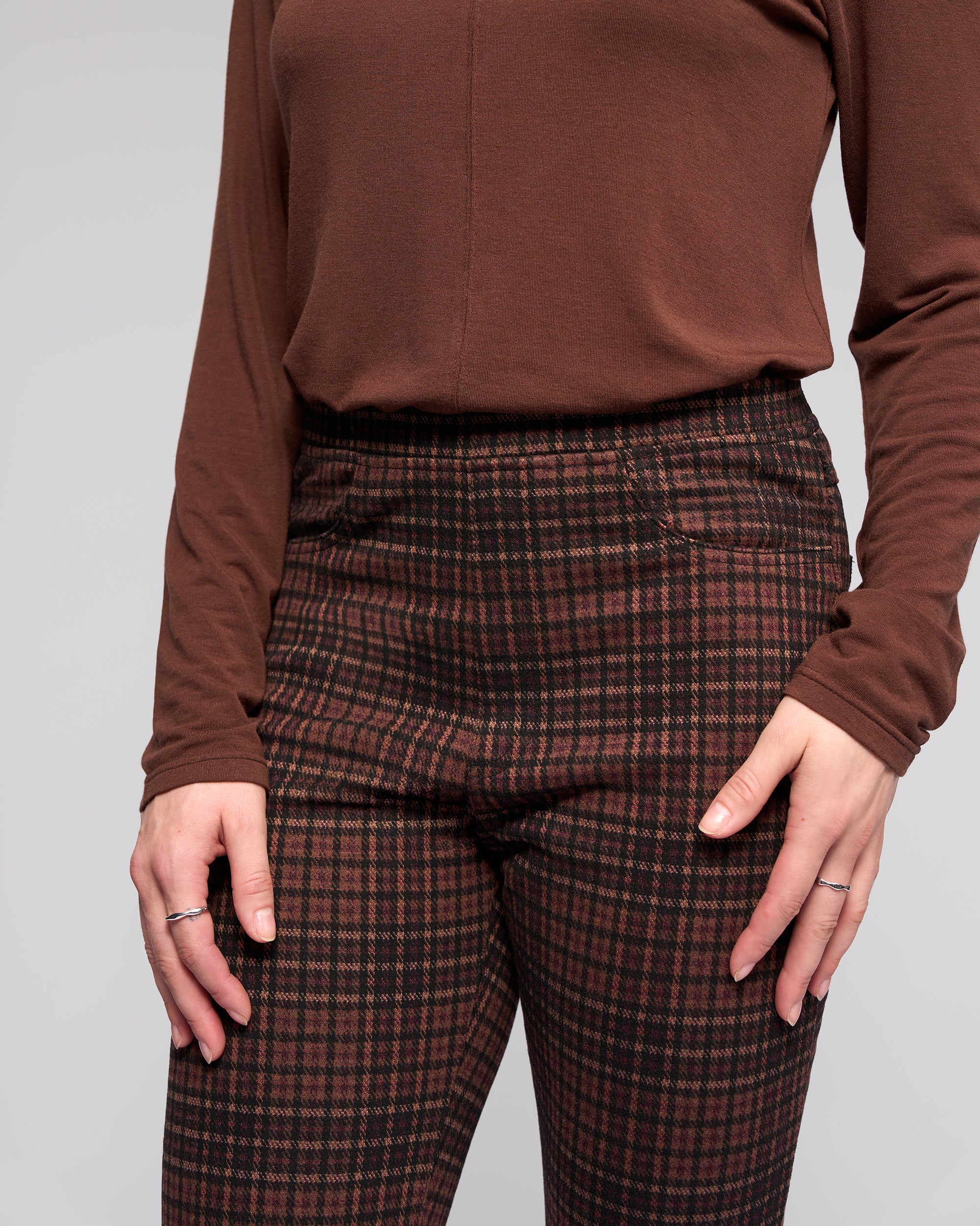 A person wears the Vassalli Bay Pull On - Cherry slim plaid pants with an elasticated waistband in brown and red, paired with a long-sleeved brown top. Thin rings adorn their hands as they rest by their sides against a plain, light background.