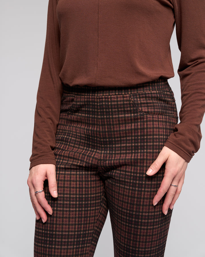 A person wears the Vassalli Bay Pull On - Cherry slim plaid pants with an elasticated waistband in brown and red, paired with a long-sleeved brown top. Thin rings adorn their hands as they rest by their sides against a plain, light background.