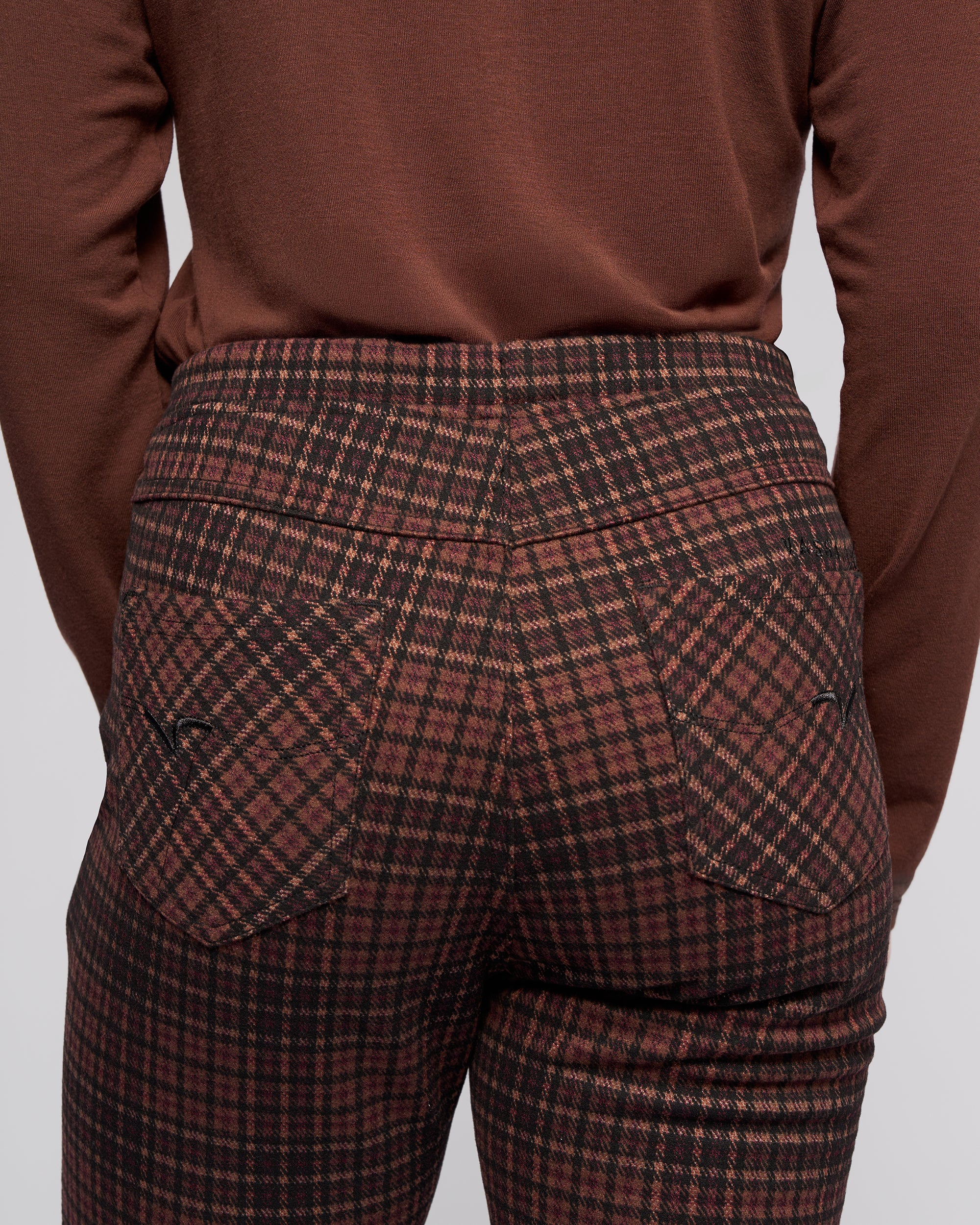A person seen from the back wears Vassalli's Bay Pull On - Cherry slim pants with an elastic waistband and pockets, paired with a brown long-sleeve shirt. The focus is on the waistband and pocket details of the pants.