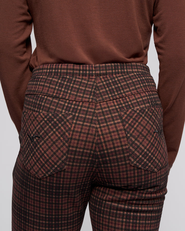 A person seen from the back wears Vassalli's Bay Pull On - Cherry slim pants with an elastic waistband and pockets, paired with a brown long-sleeve shirt. The focus is on the waistband and pocket details of the pants.