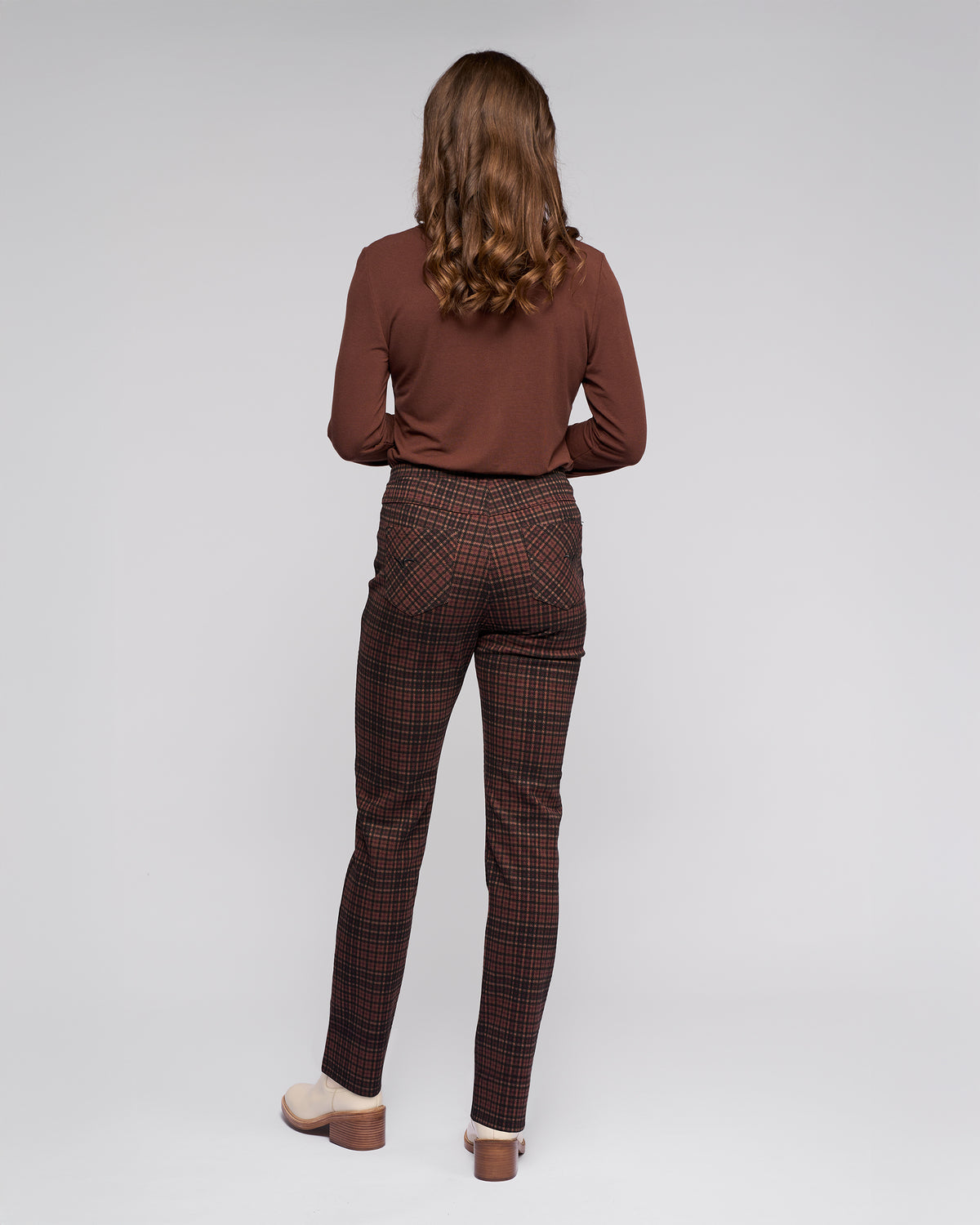 A person with wavy brown hair, facing away, wears a long-sleeve brown top and Vassalli Bay Pull On - Cherry plaid slim pants with an elasticated waistband and light shoes, standing against a plain gray background.