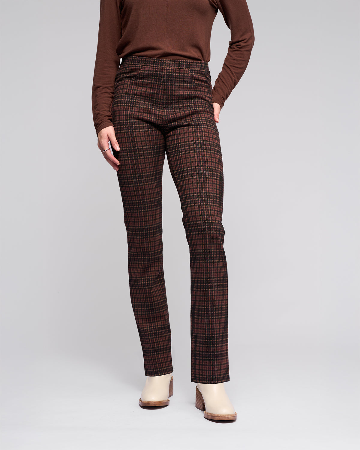 A person wears Vassalli Bay Pull On - Cherry pants with an elasticated waistband, a brown long-sleeved top, and beige ankle boots, standing against a plain light gray background; their face is not visible.