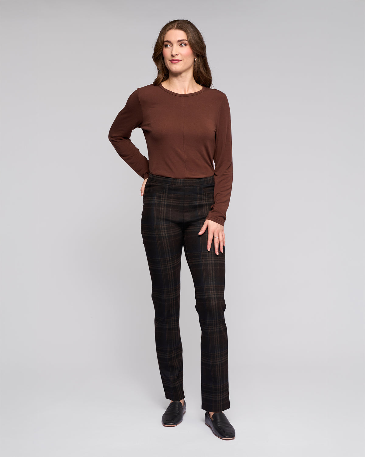 A woman stands against a plain background wearing the Vassalli Bay Pull On - Hugo dark slim pants with an elasticated waistband in stretch ponti fabric, a long-sleeve brown top, and black slip-on shoes. She has long brown hair and is smiling slightly.