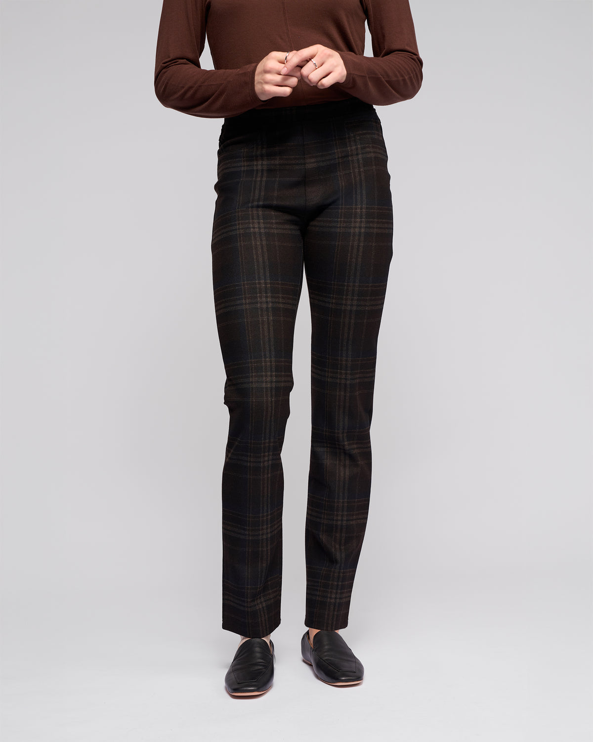 Person wearing Vassalli’s Bay Pull On - Hugo, a high-waisted dark plaid slim pant with elastic waistband, paired with a long-sleeved brown top. Stands against a light gray background; only the body from shoulders down is visible.