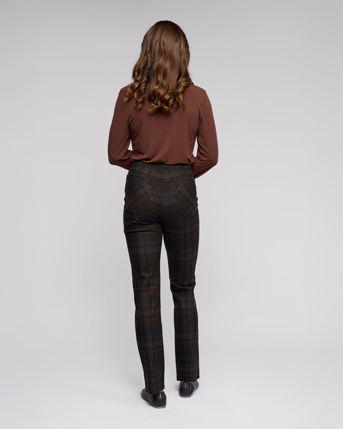 A person with long, wavy brown hair, facing away from the camera, wears a brown long-sleeve shirt with Vassalli’s Bay Pull On - Hugo plaid slim pants and black shoes against a plain light gray background.