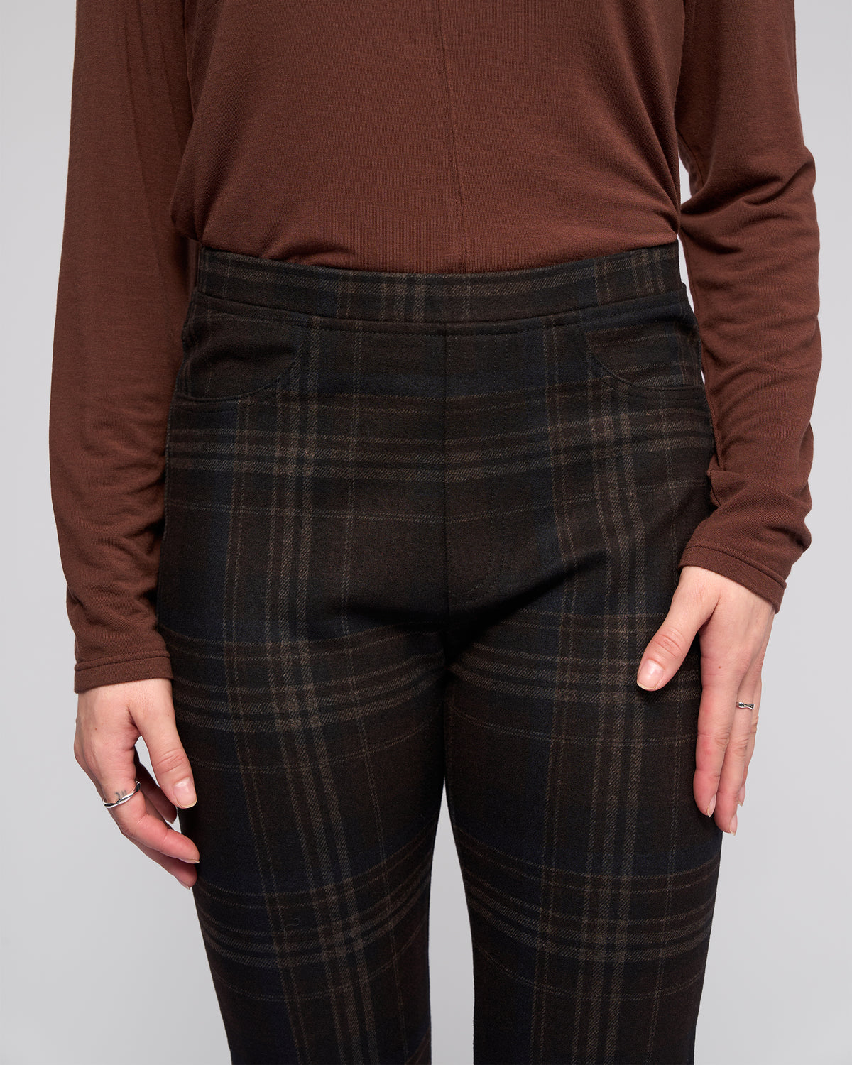 A person showcases the Vassalli Bay Pull On - Hugo black plaid slim pant with an elasticated waistband, paired with a long-sleeve brown top, standing with arms relaxed by their sides against a plain light background.