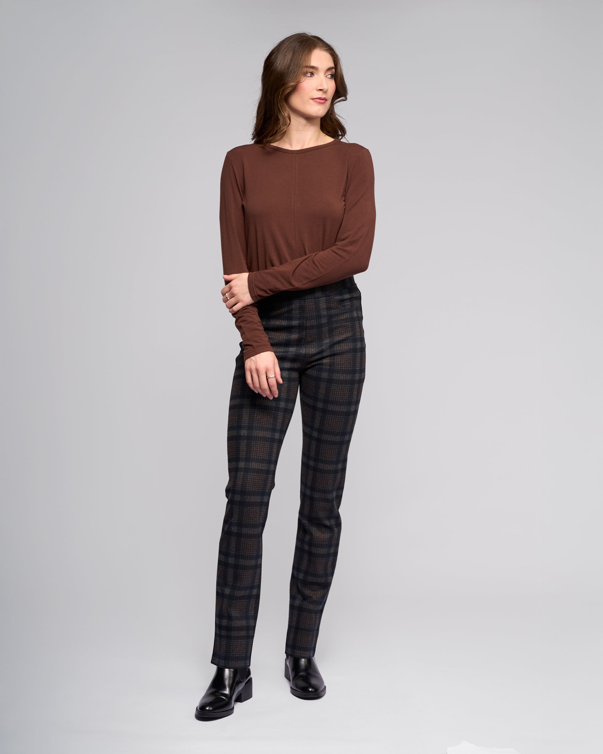 A woman with wavy brown hair stands in a studio wearing Vassalli Bay Pull On - Nomad slim plaid stretch ponti trousers, a long-sleeve brown top, and black shoes, looking to the side with one arm across her body against a plain light background.
