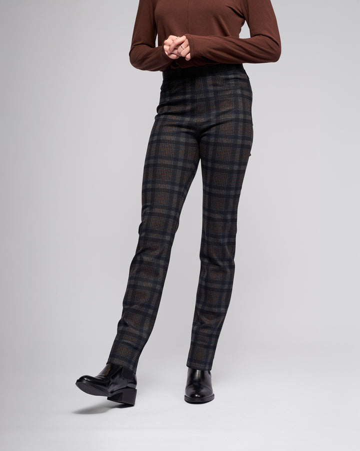 A person models Vassalli's Bay Pull On - Nomad slim stretch ponti pants in dark plaid with a brown long-sleeve top and black heels against a plain light gray background, shown from the shoulders down.