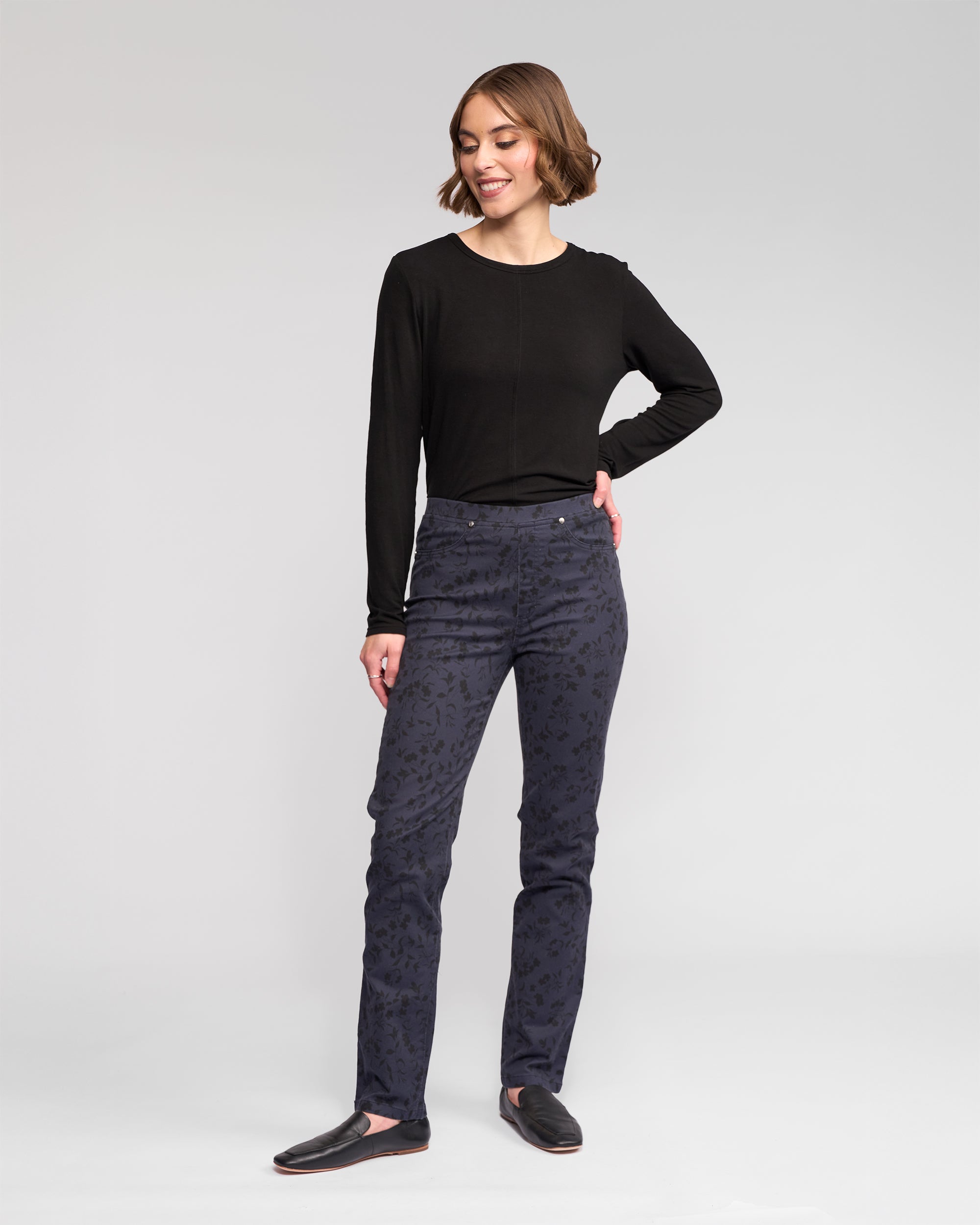 A woman stands against a plain background, smiling and looking slightly to the side. She wears black long-sleeve top, Vassalli Bay Pull On - Noir slim stretch printed denim pants, and black slip-on shoes, with one hand on her hip.