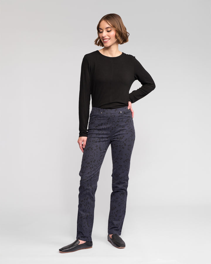A woman stands against a plain background, smiling and looking slightly to the side. She wears black long-sleeve top, Vassalli Bay Pull On - Noir slim stretch printed denim pants, and black slip-on shoes, with one hand on her hip.