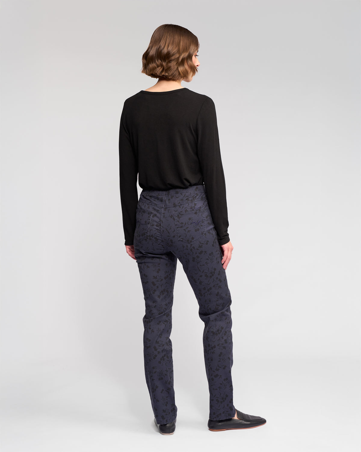 A person with short brown hair faces away, wearing a long-sleeved black top, black shoes, and Vassalli Bay Pull On - Noir pants, against a plain light background.