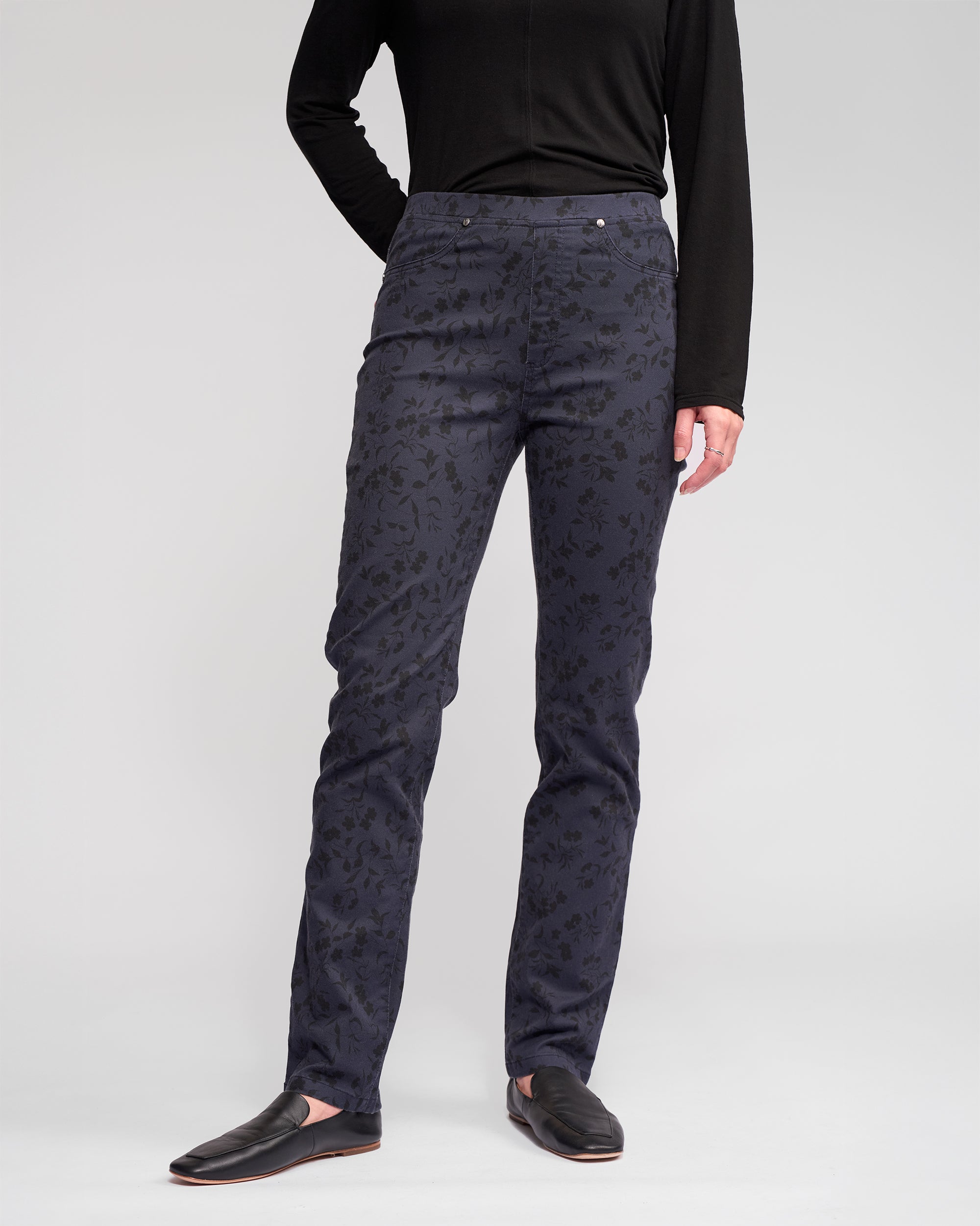 A person, face not visible, stands against a plain light gray background wearing a black long-sleeve top, Vassalli Bay Pull On - Noir slim black floral pants, and black slip-on shoes.