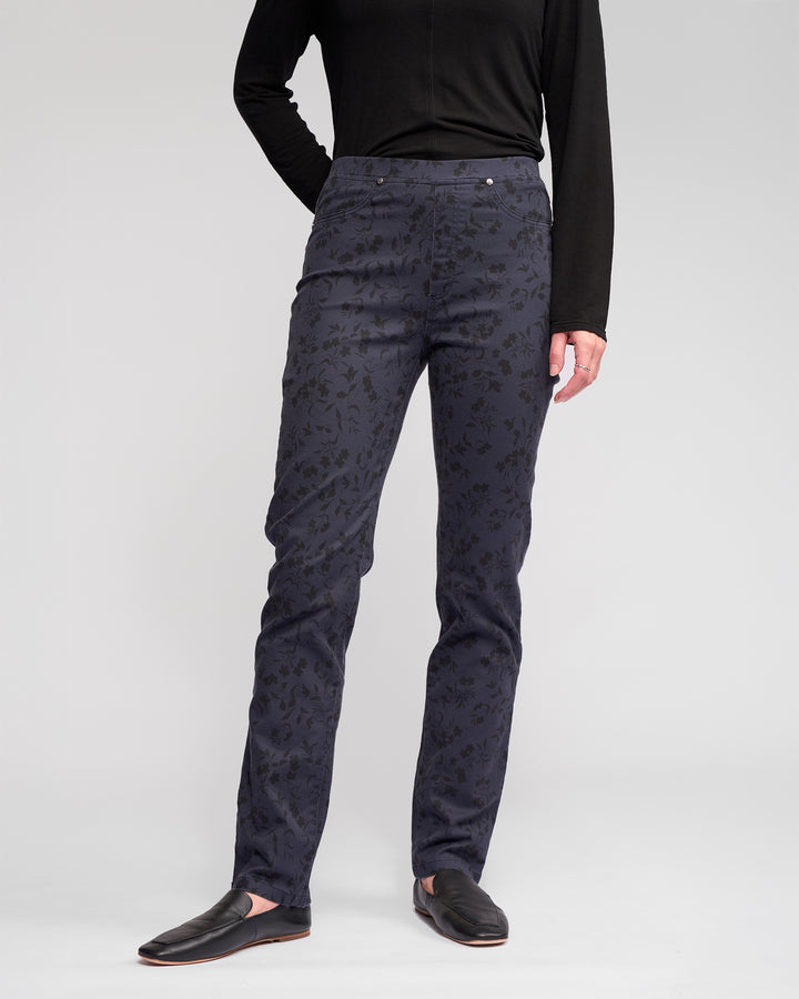A person, face not visible, stands against a plain light gray background wearing a black long-sleeve top, Vassalli Bay Pull On - Noir slim black floral pants, and black slip-on shoes.