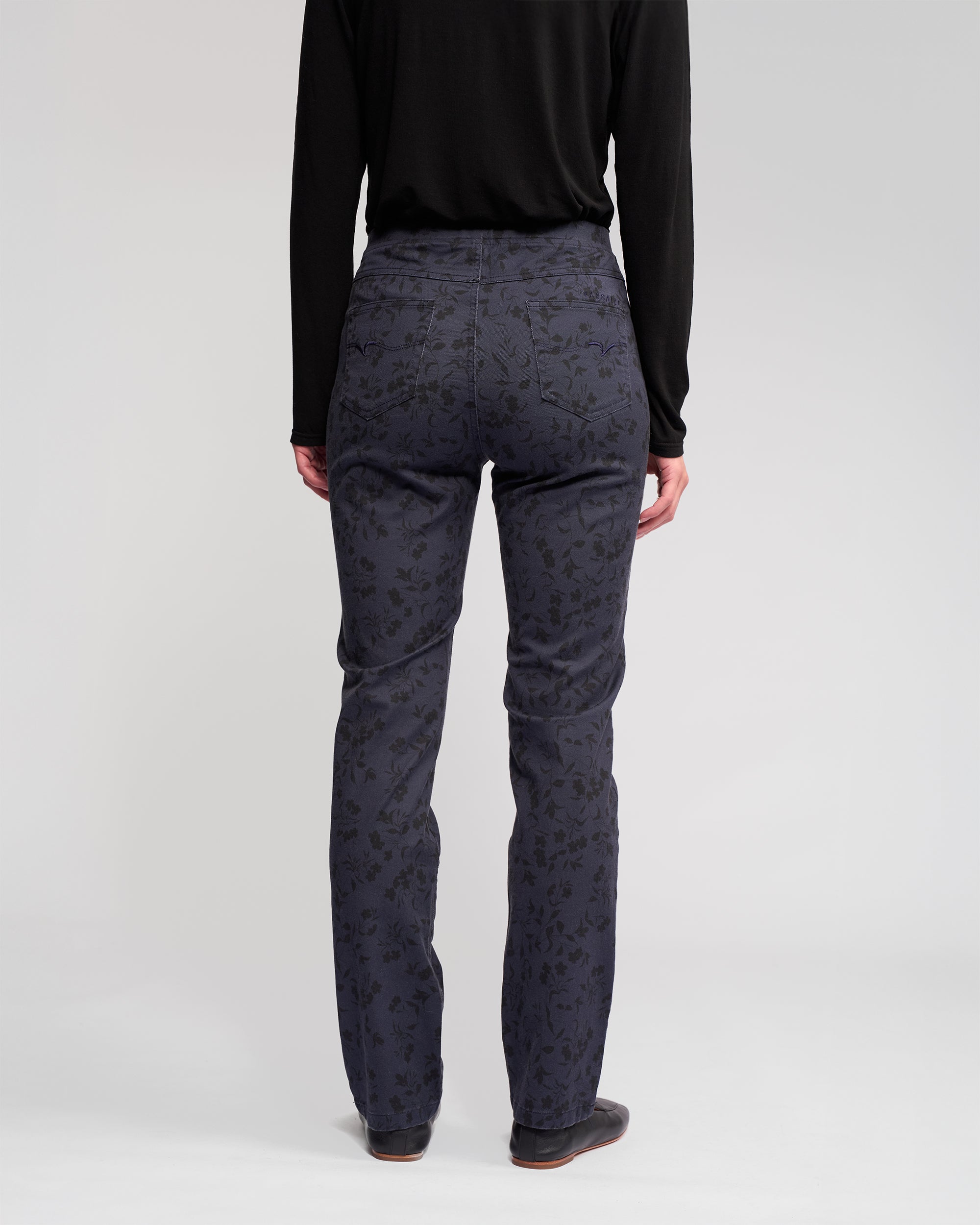 A person stands facing away, wearing Vassalli’s Bay Pull On - Noir slim pant in dark blue with a subtle floral pattern, paired with a black long-sleeve top and dark shoes against a plain light background.