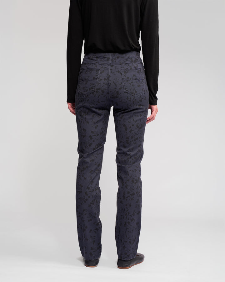 A person stands facing away, wearing Vassalli’s Bay Pull On - Noir slim pant in dark blue with a subtle floral pattern, paired with a black long-sleeve top and dark shoes against a plain light background.