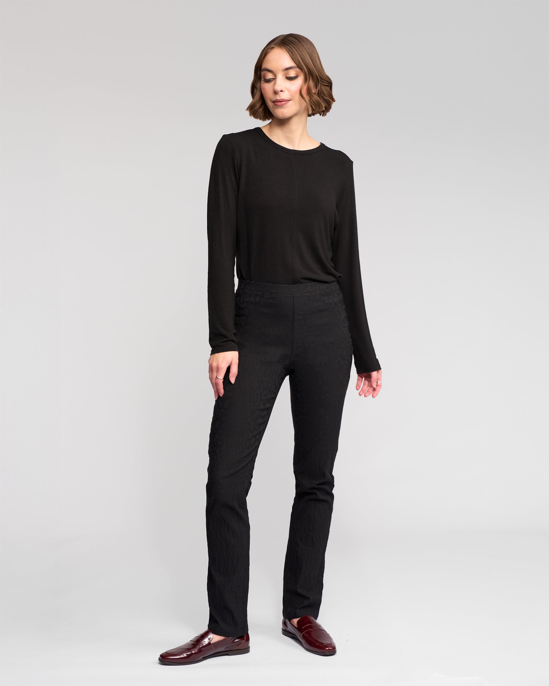 A woman with short brown hair stands against a plain background, wearing Vassalli's Bay Pull On - Black pants and a long-sleeve black top, paired with dark red loafers. She looks slightly to the side with a relaxed expression.