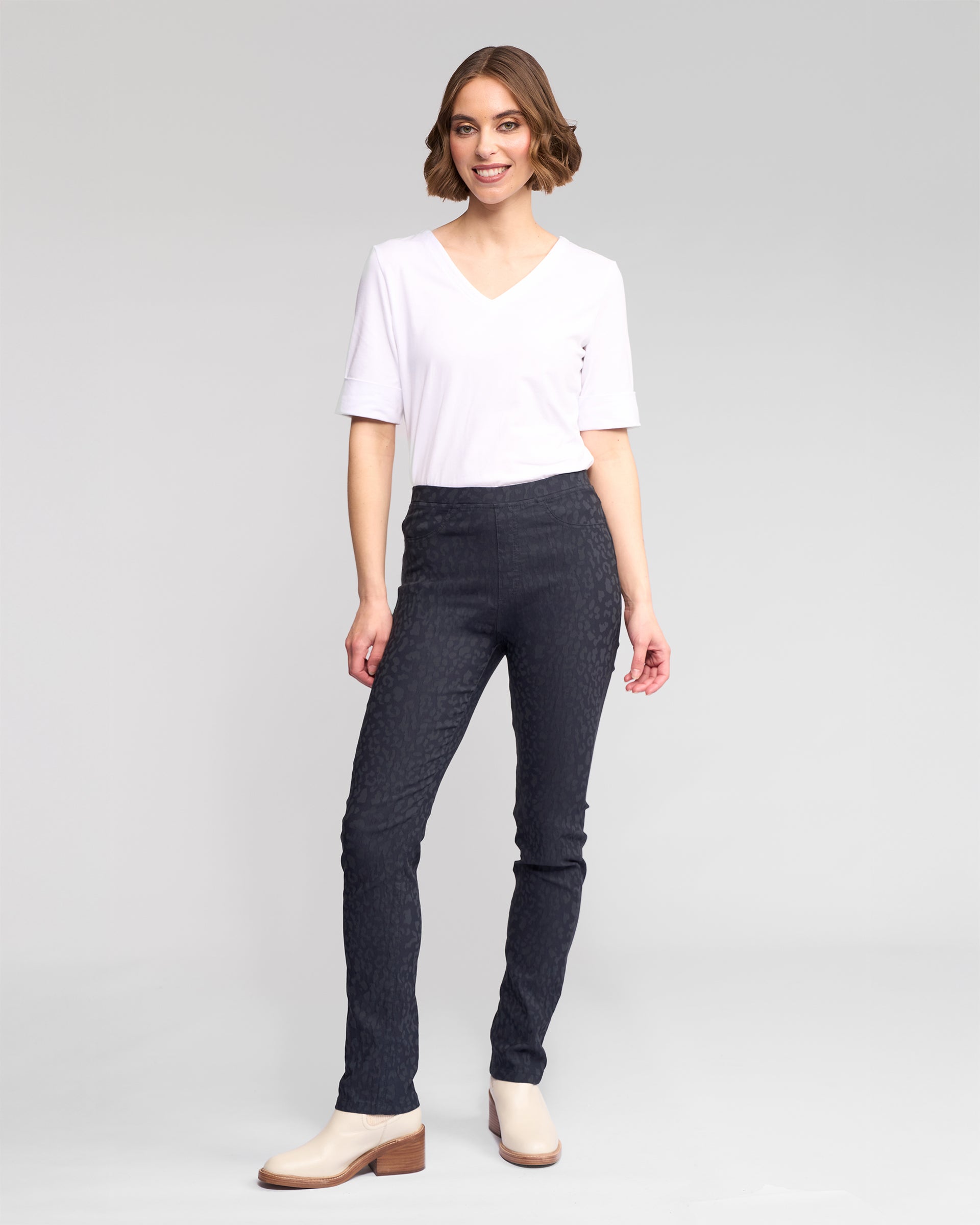 A woman with short brown hair, dressed in a white V-neck T-shirt, light ankle boots, and Vassalli Bay Pull On - Navy slim animal-textured pants with an elastic waist, smiles against a plain light gray background.