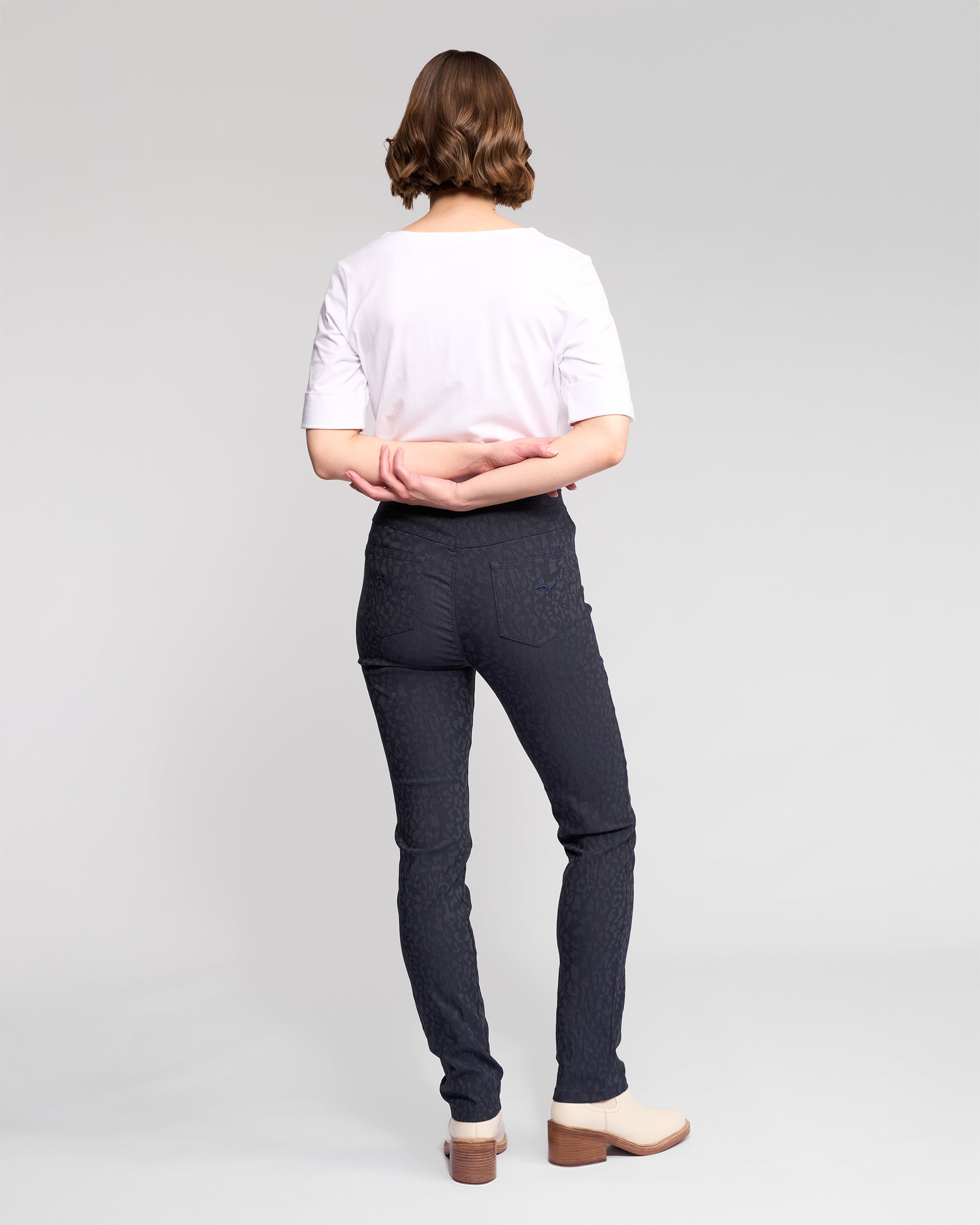 A person with short wavy hair stands facing away in a white t-shirt and Vassalli Bay Pull On - Navy pants with an elastic waistband, paired with light boots, arms loosely crossed behind them against a plain light background.