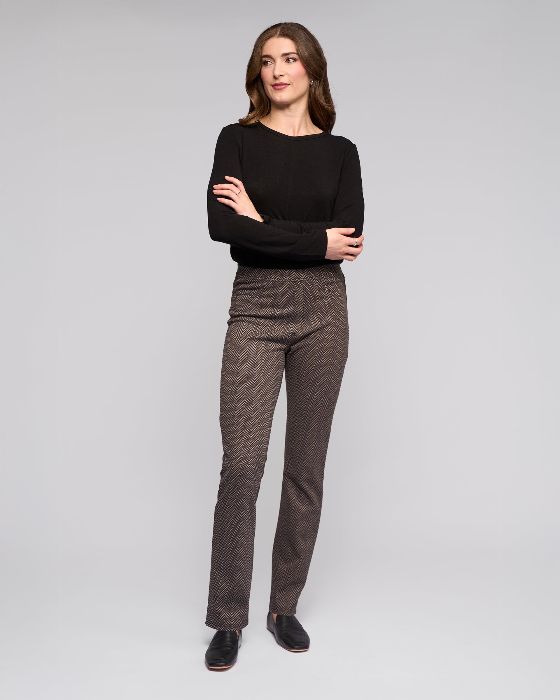 A woman with wavy brown hair stands against a light gray background, wearing Vassalli Bay Pull On - Arc patterned brown slim pants and a black long-sleeve top. She smiles slightly, arms crossed, and completes the look with black shoes.