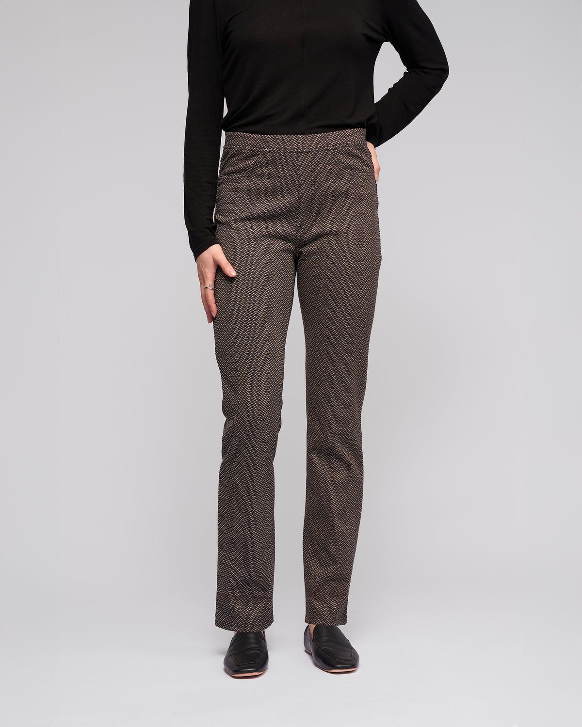Someone stands against a plain light background wearing a black long-sleeve top, black shoes, and Vassalli Bay Pull On - Arc patterned brown slim pants; their right hand rests on their hip.