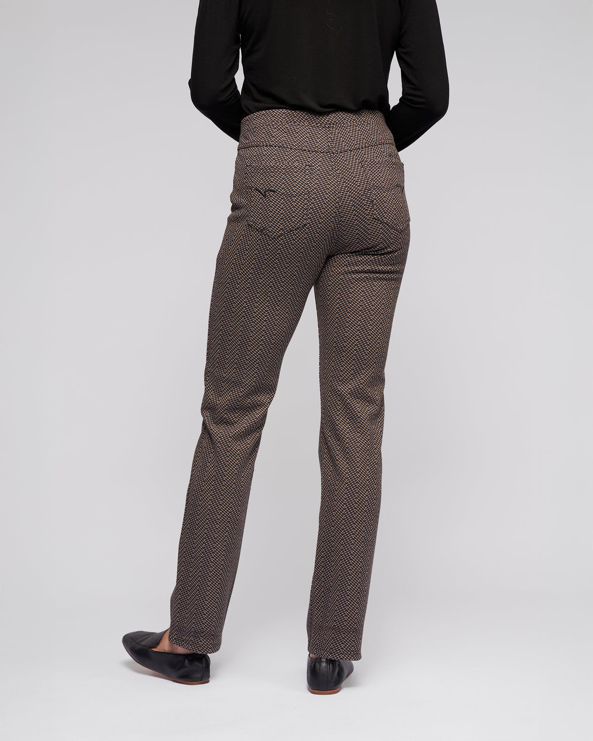 A person faces away, wearing Vassalli Bay Pull On - Arc brown herringbone pants, a black long-sleeve top, and black shoes against a plain light gray background.