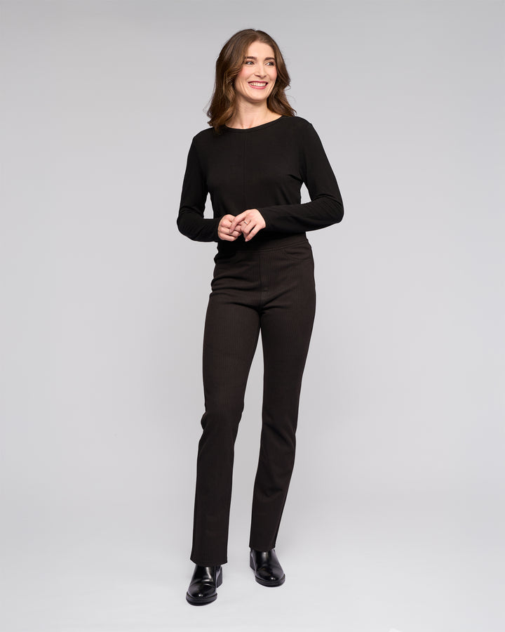 A woman with wavy brown hair smiles against a light gray background, wearing a black long-sleeve top and Vassalli Bay Pull On - Metro slim knit pants, styled with black ankle boots.