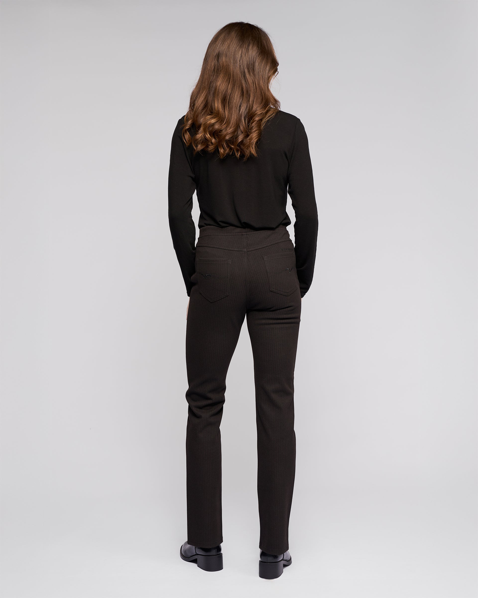 A person with long, wavy brown hair stands facing away from the camera wearing a Vassalli Bay Pull On - Metro slim pant, a black top, and black shoes against a plain light background.