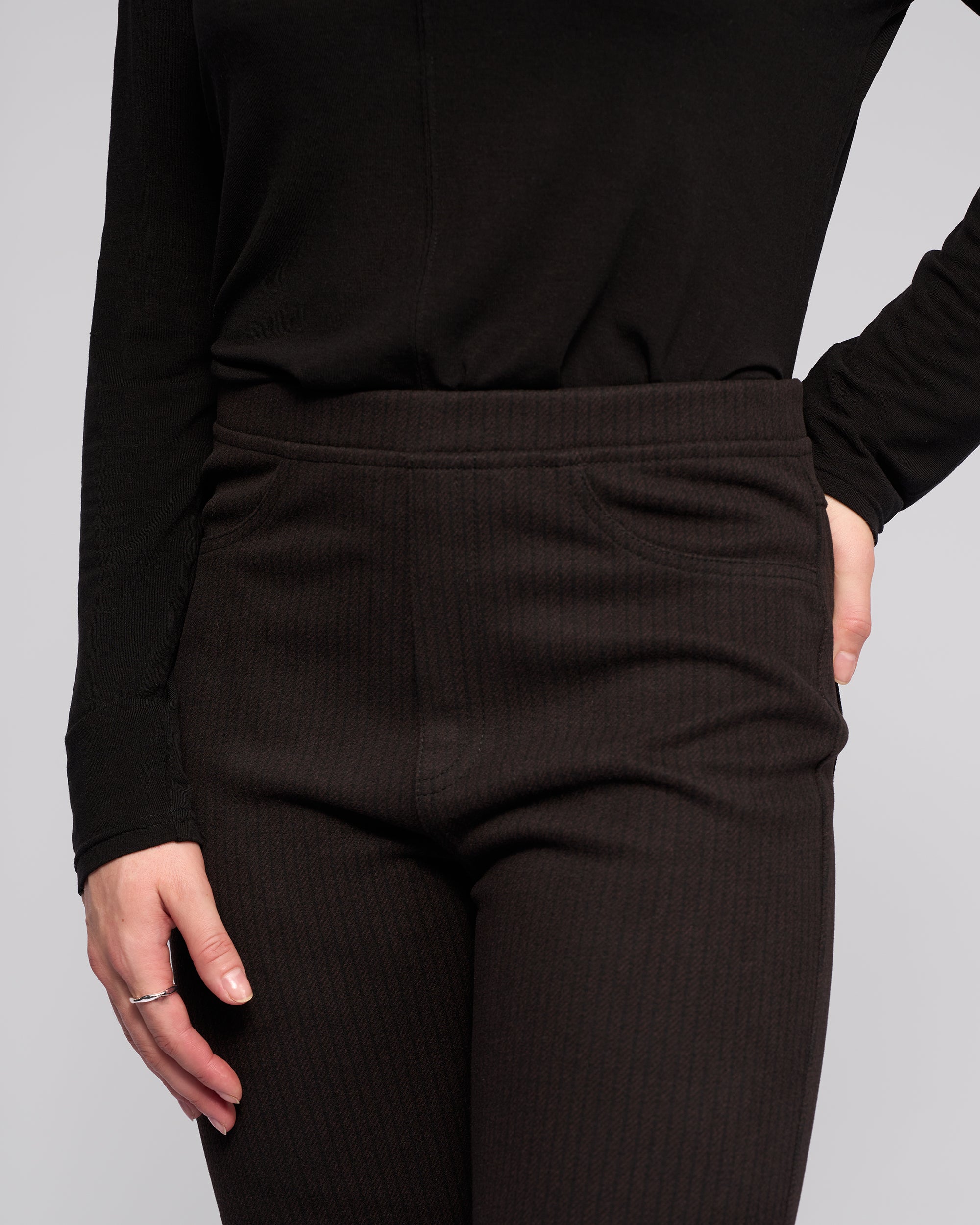 A person in a long-sleeve black top and Vassalli Bay Pull On - Metro slim knit pants stands with one hand in their pocket and the other by their side, shown from shoulders to mid-thigh.