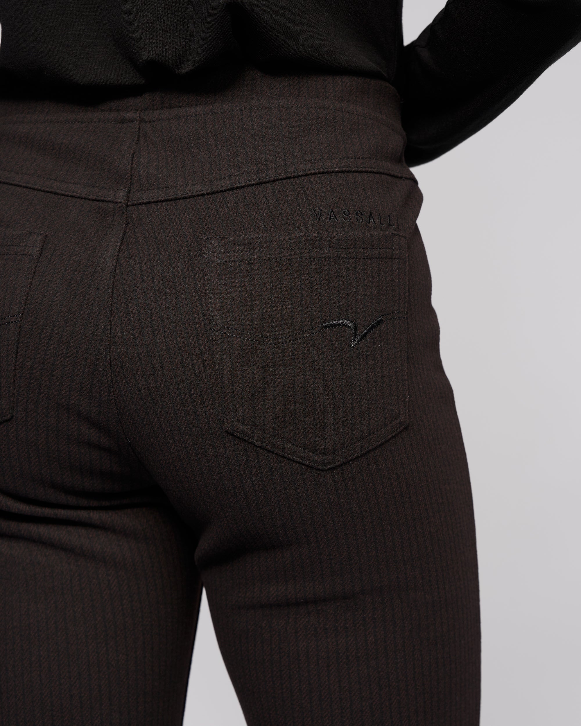 Close-up of a person wearing Vassalli Bay Pull On - Metro dark, pinstriped slim pants and a black top, featuring the back pocket with the brand name stitched above it.