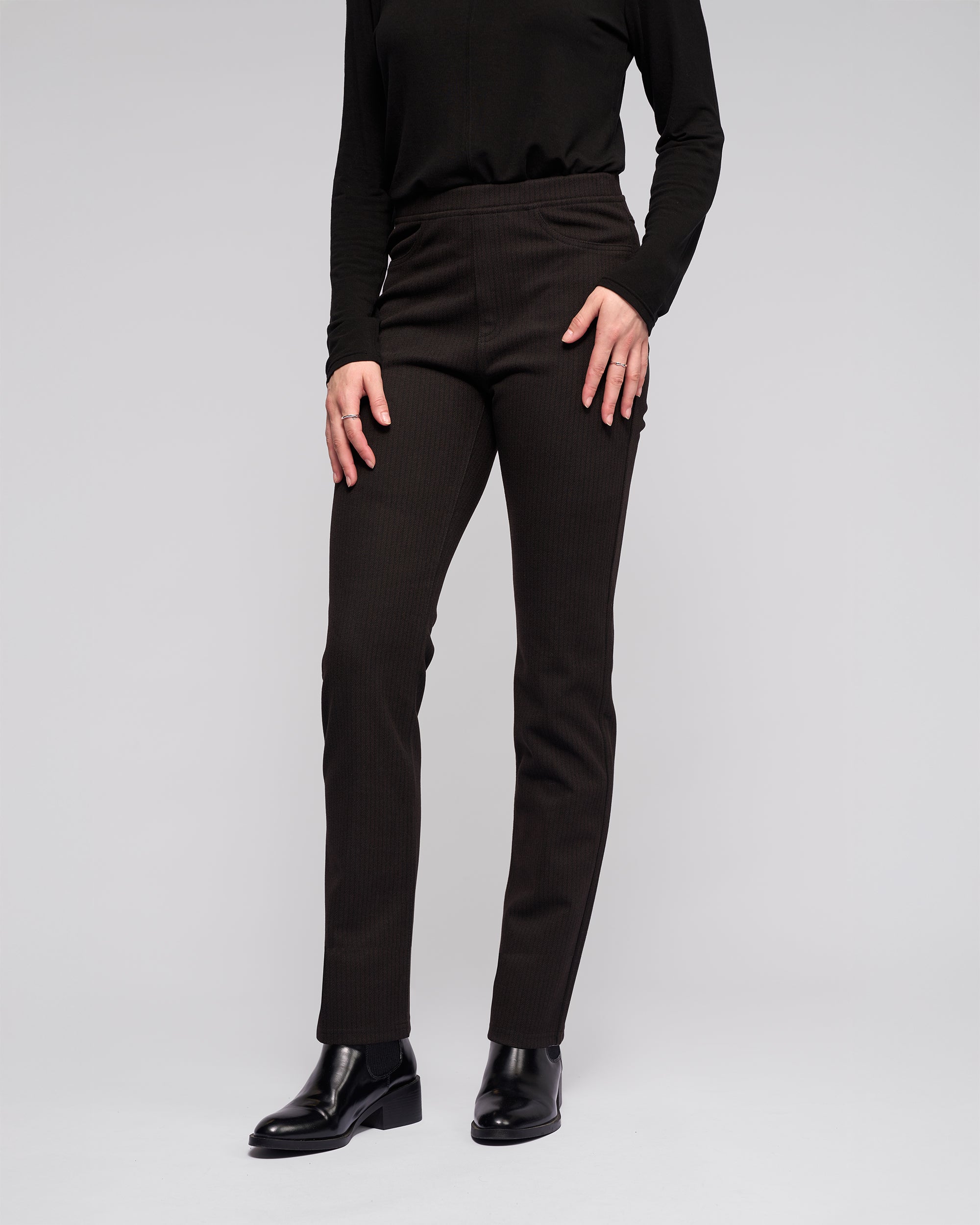 A person wearing Vassalli's Bay Pull On - Metro fitted black slim pants, a black long-sleeve top, and black ankle boots stands against a plain light gray background. The face is not visible.