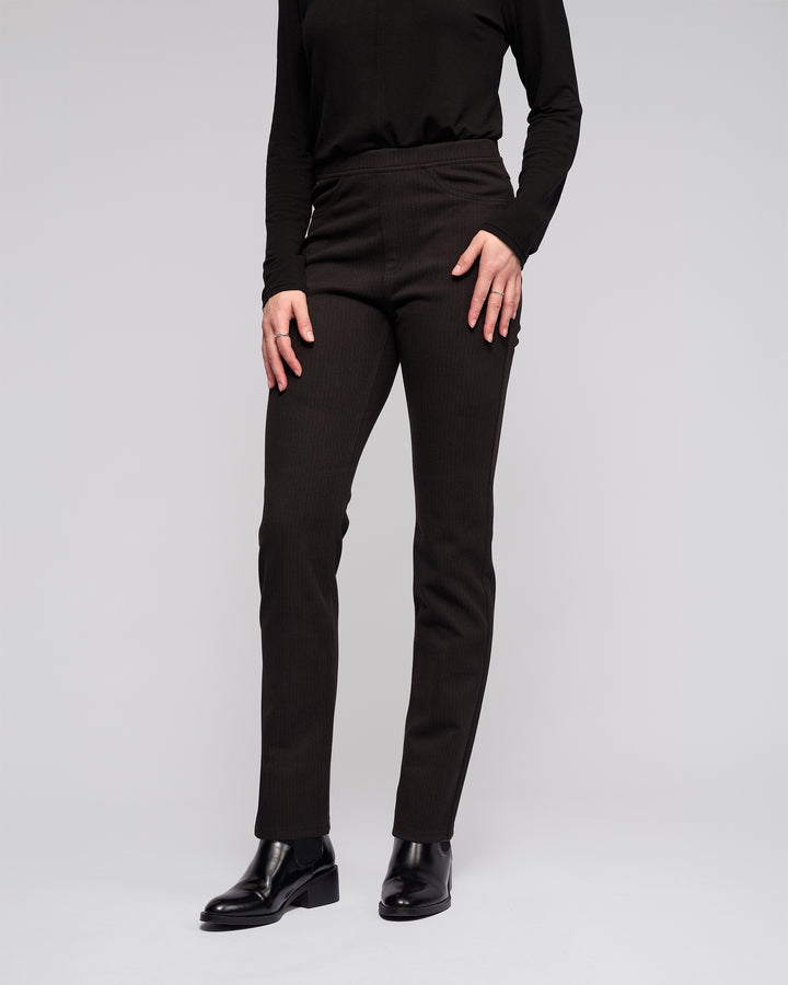 A person wearing Vassalli's Bay Pull On - Metro fitted black slim pants, a black long-sleeve top, and black ankle boots stands against a plain light gray background. The face is not visible.