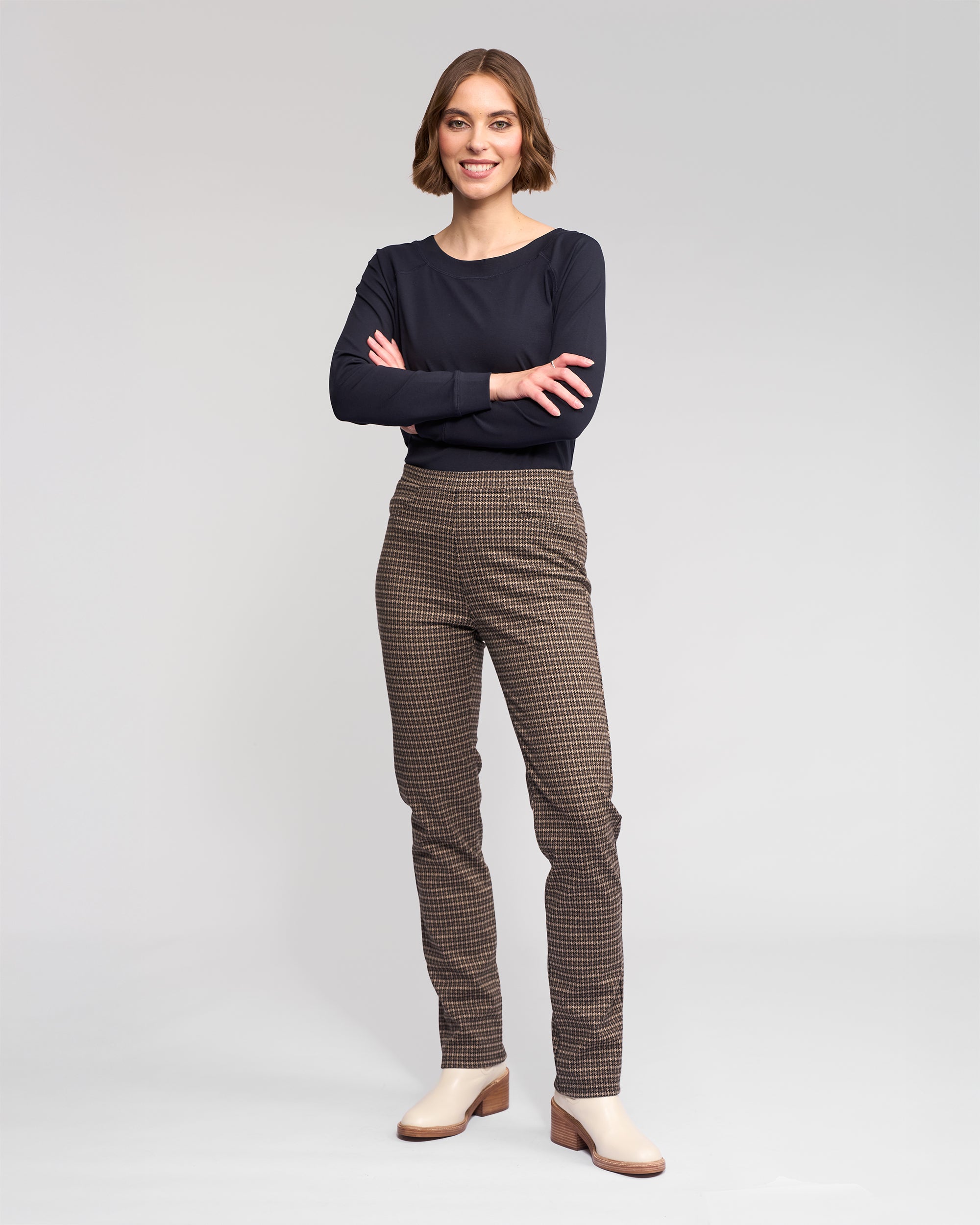 A woman with short brown hair smiles with arms crossed, wearing Vassalli Bay Pull On - Nova brown checkered slim pants in comfy knit, a black long-sleeve top, and beige heels against a plain light gray background.
