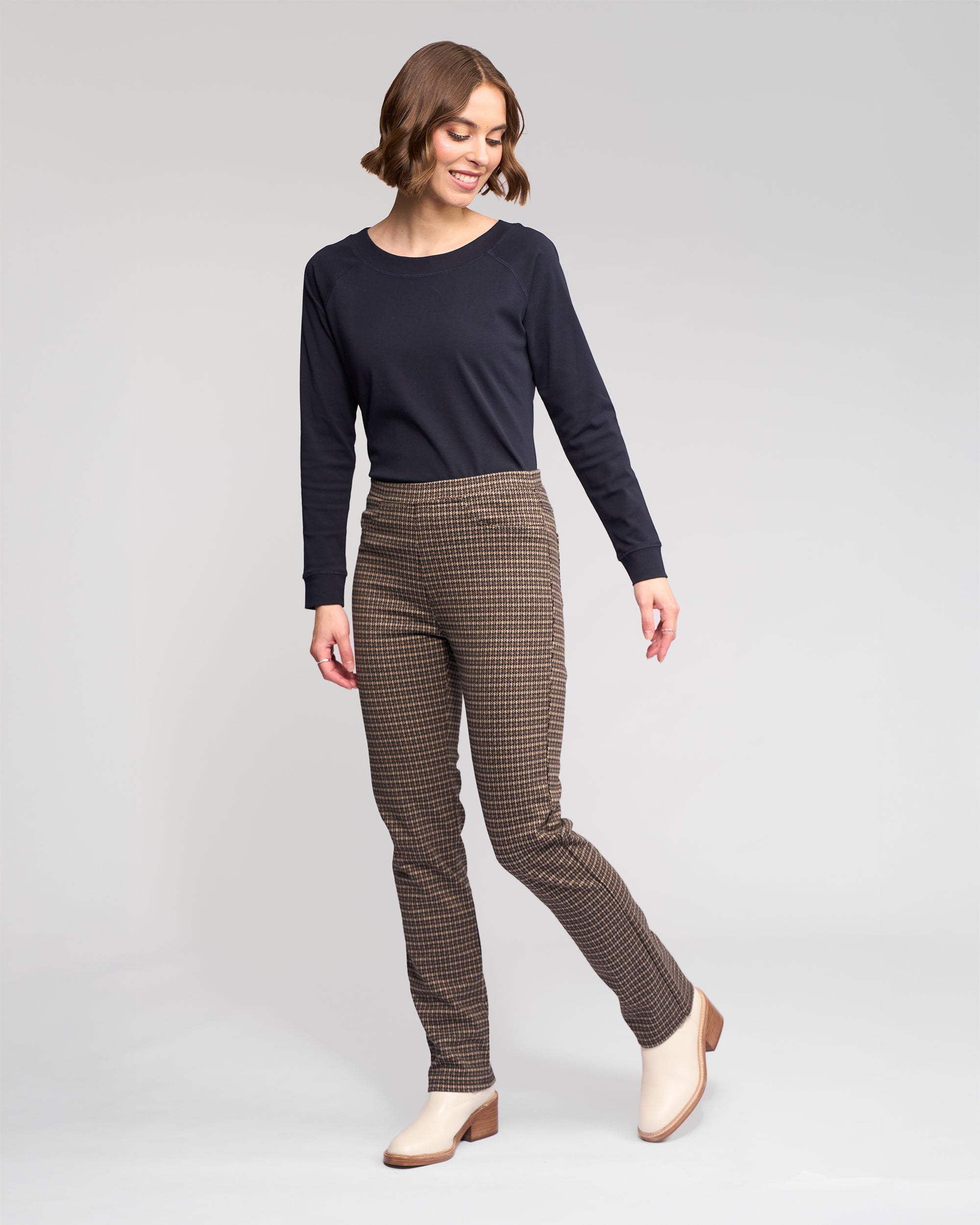 A woman with short brown hair smiles and looks down, dressed in Vassalli's Bay Pull On - Nova brown plaid slim pants and a long-sleeve black top, paired with beige heeled shoes against a plain light background.