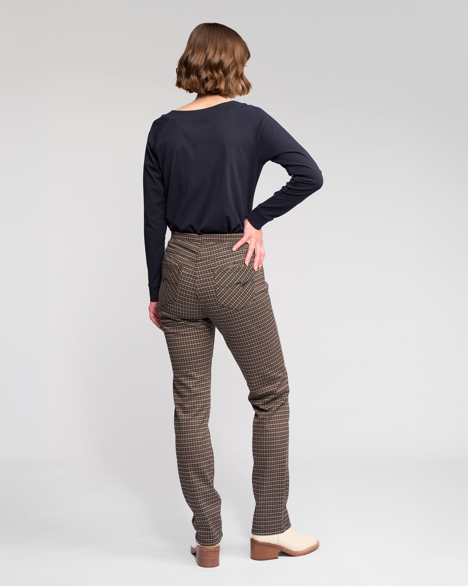 Wearing the Vassalli Bay Pull On - Nova brown slim knit pants, a person with short brown hair and a black long-sleeve top poses in beige heeled boots, standing against a plain light background with one hand on their hip.
