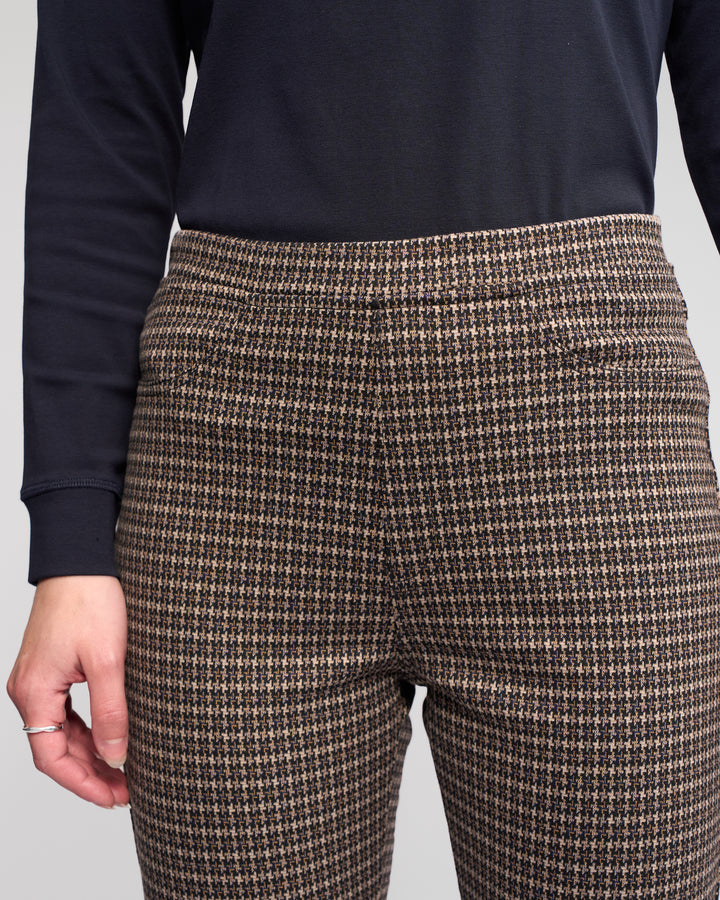 A person wearing the Vassalli Bay Pull On - Nova, high-waisted brown and black houndstooth slim pants in comfortable knit, paired with a dark long-sleeve shirt, their left hand resting at their side.