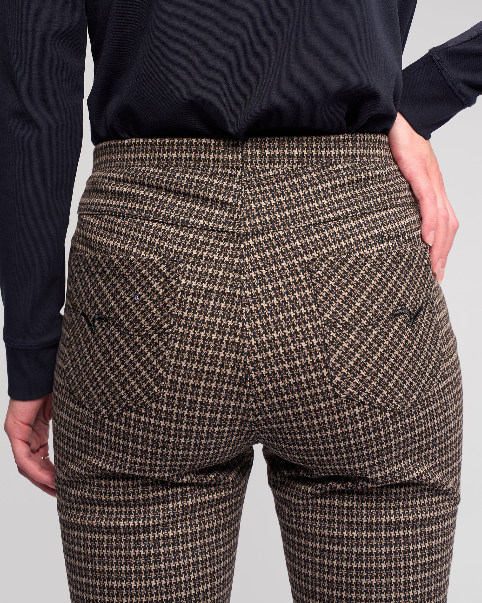 Someone is shown from the back, hands relaxed by their sides, wearing Vassalli's Bay Pull On - Nova high-waisted slim pants in brown and black houndstooth knit with a black long-sleeve top.