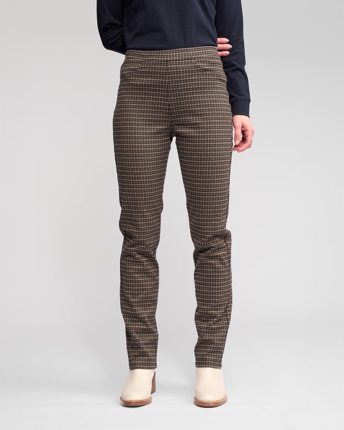 A person models Vassalli's Bay Pull On - Nova brown checkered slim pants with a long-sleeve black top and beige ankle boots, standing against a plain light gray background. The comfortable knit fabric pants are shown cropped at the shoulders.
