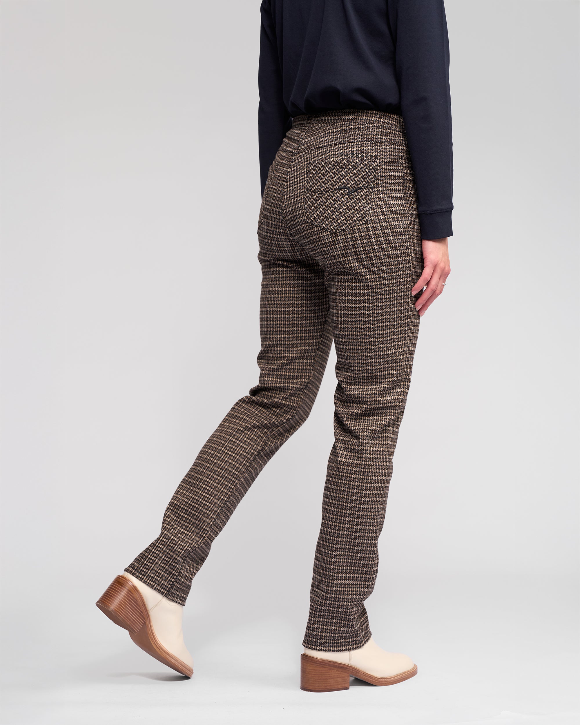 A person wearing a dark long-sleeve shirt, Vassalli Bay Pull On - Nova slim plaid pants in comfortable knit fabric, and beige heeled ankle boots stands facing away with one foot slightly raised against a plain light background.