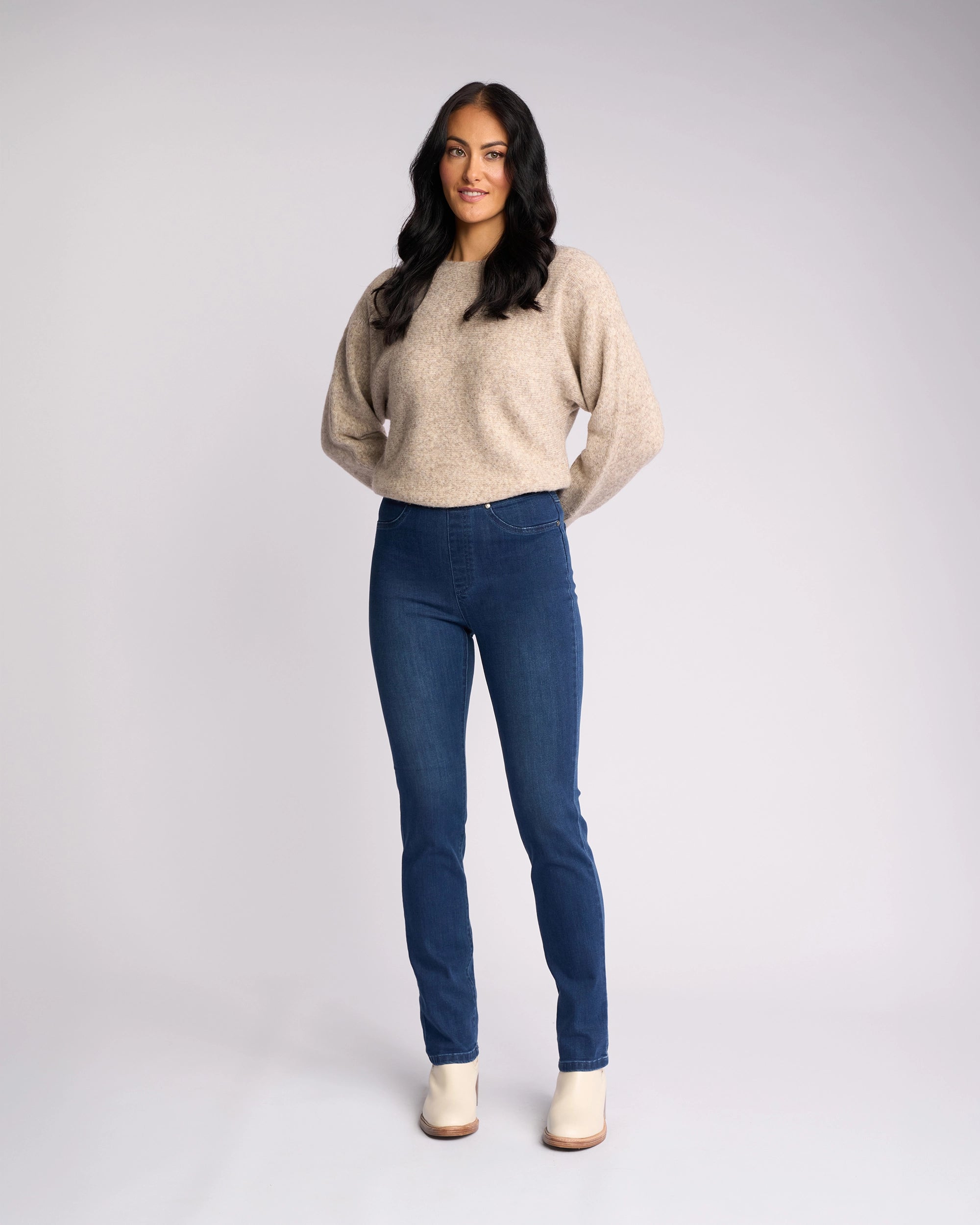 A woman with long dark hair faces forward, wearing a light beige sweater and Vassalli's Bay Pull On - Blue Denim (With Front Fade) jeans, paired with light ankle boots, against a plain light gray background.