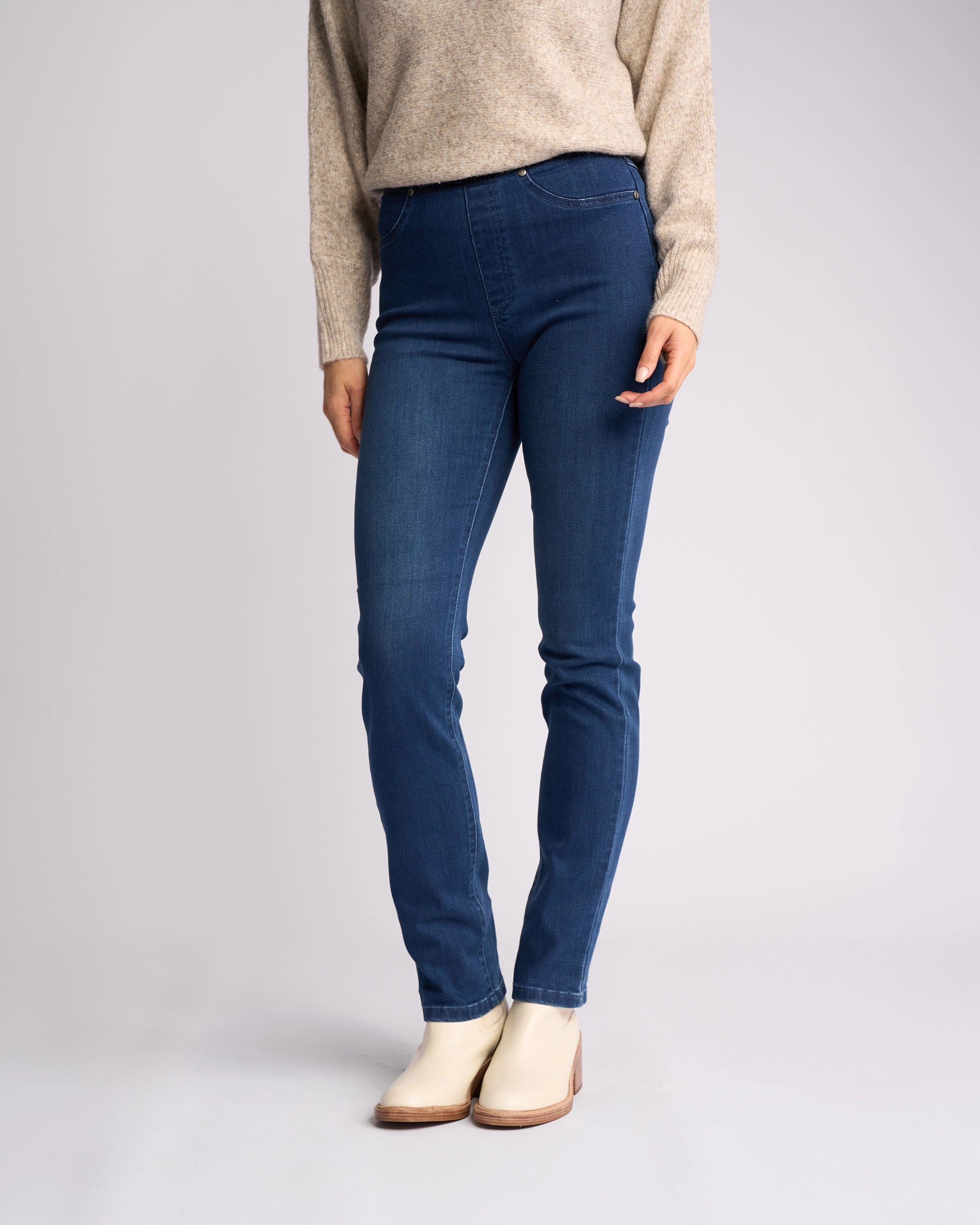 A person stands against a plain light background wearing Vassalli's Bay Pull On - Blue Denim (With Front Fade) jeans, a beige sweater, and cream ankle boots. The person's face is not visible.