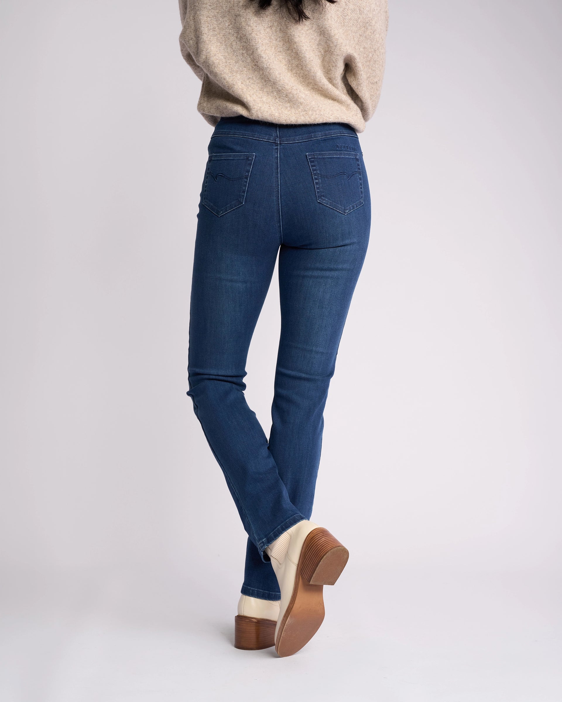 A person stands with their back to the camera, wearing Vassalli's Bay Pull On - Blue Denim (With Front Fade) jeans, a beige sweater, and light heeled shoes against a plain light background.