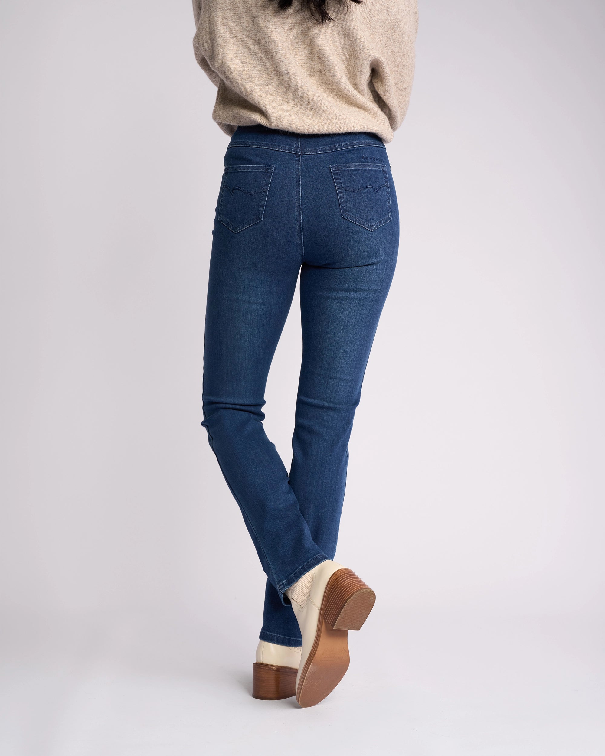 A person stands with their back to the camera, wearing Vassalli's Bay Pull On - Blue Denim (With Front Fade) jeans, a beige sweater, and light heeled shoes against a plain light background.