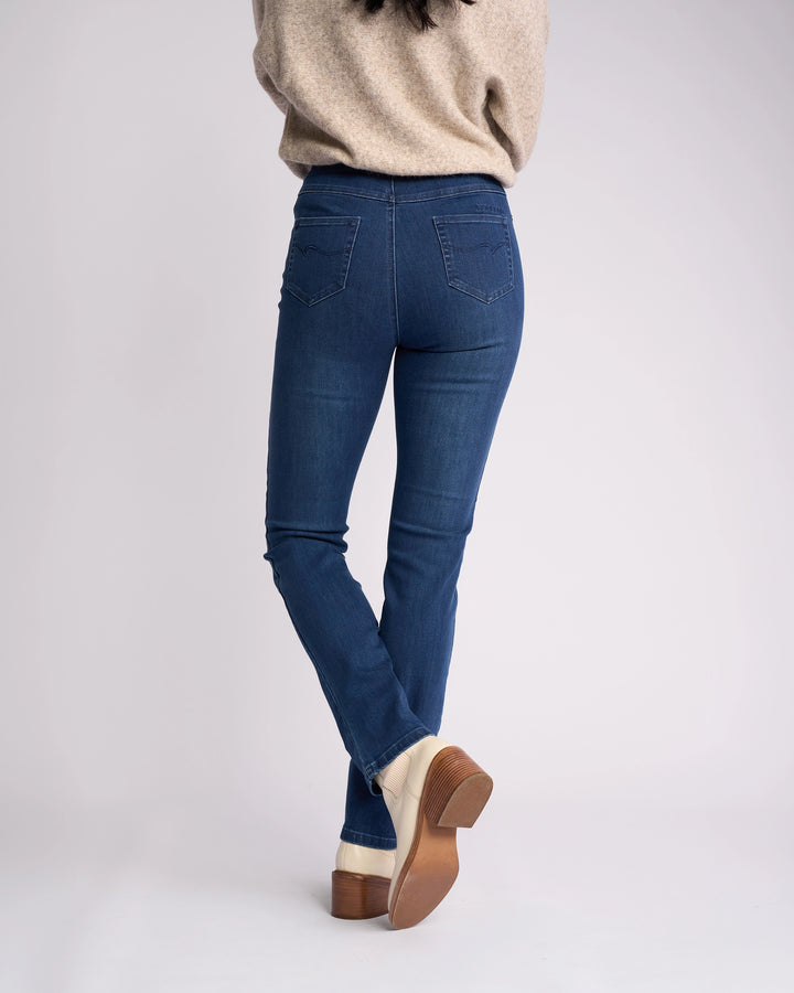 A person stands with their back to the camera, wearing Vassalli's Bay Pull On - Blue Denim (With Front Fade) jeans, a beige sweater, and light heeled shoes against a plain light background.