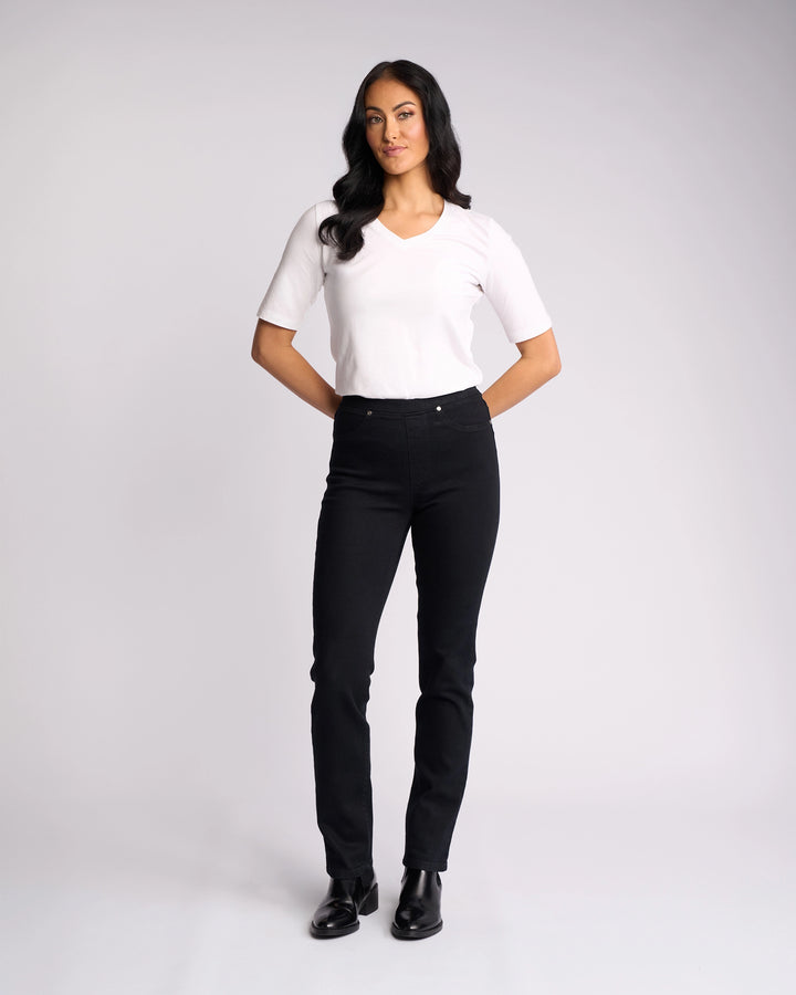 A woman with long dark hair stands against a light background, wearing a white short-sleeve shirt, black shoes, and Vassalli Bay Pull On - Black Denim slim-leg jeans with her hands clasped behind her back.
