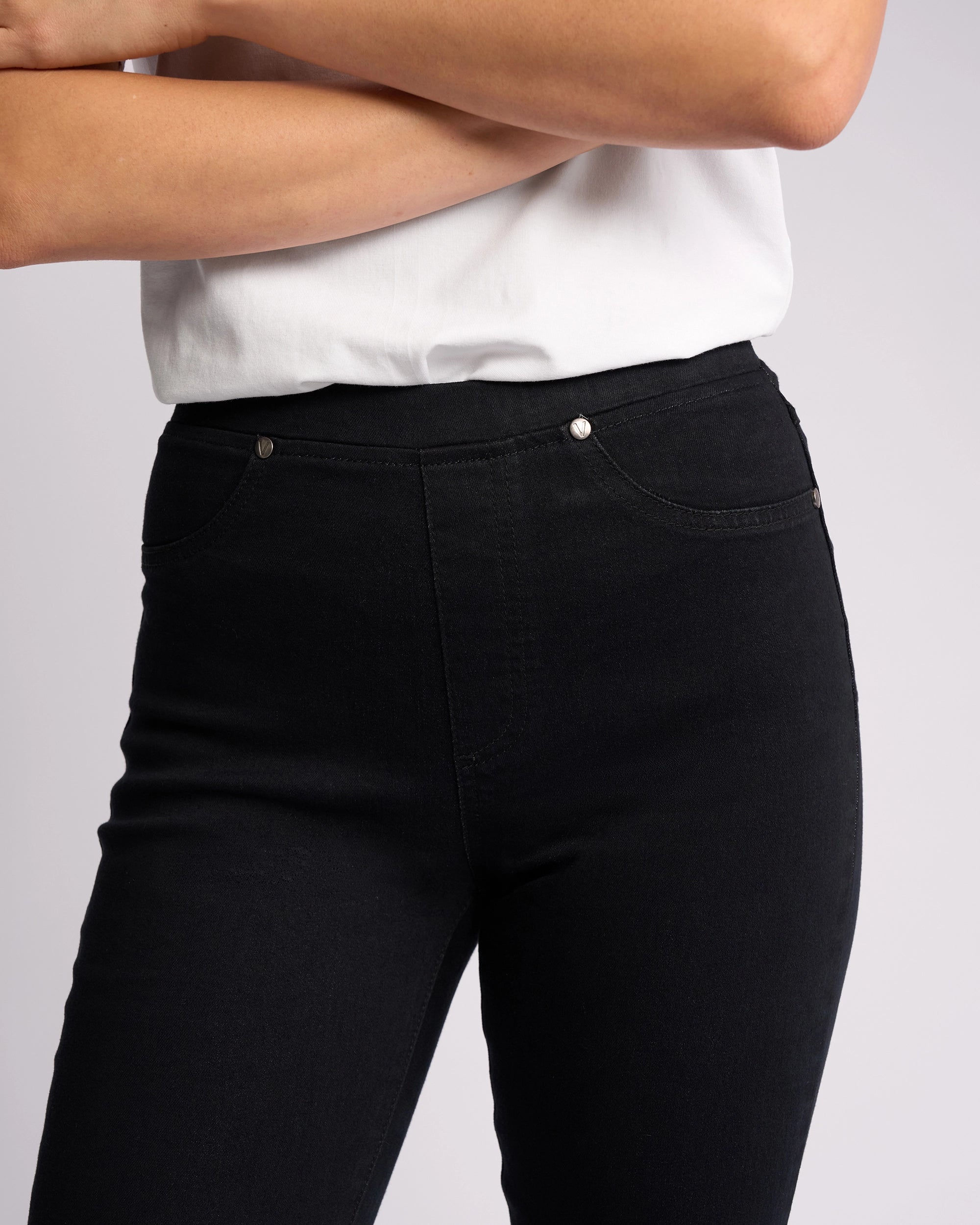 A person wearing a white short-sleeve shirt and Vassalli Bay Pull On - Black Denim slim-leg jeans, arms crossed, shown from shoulders to thighs against a plain background.