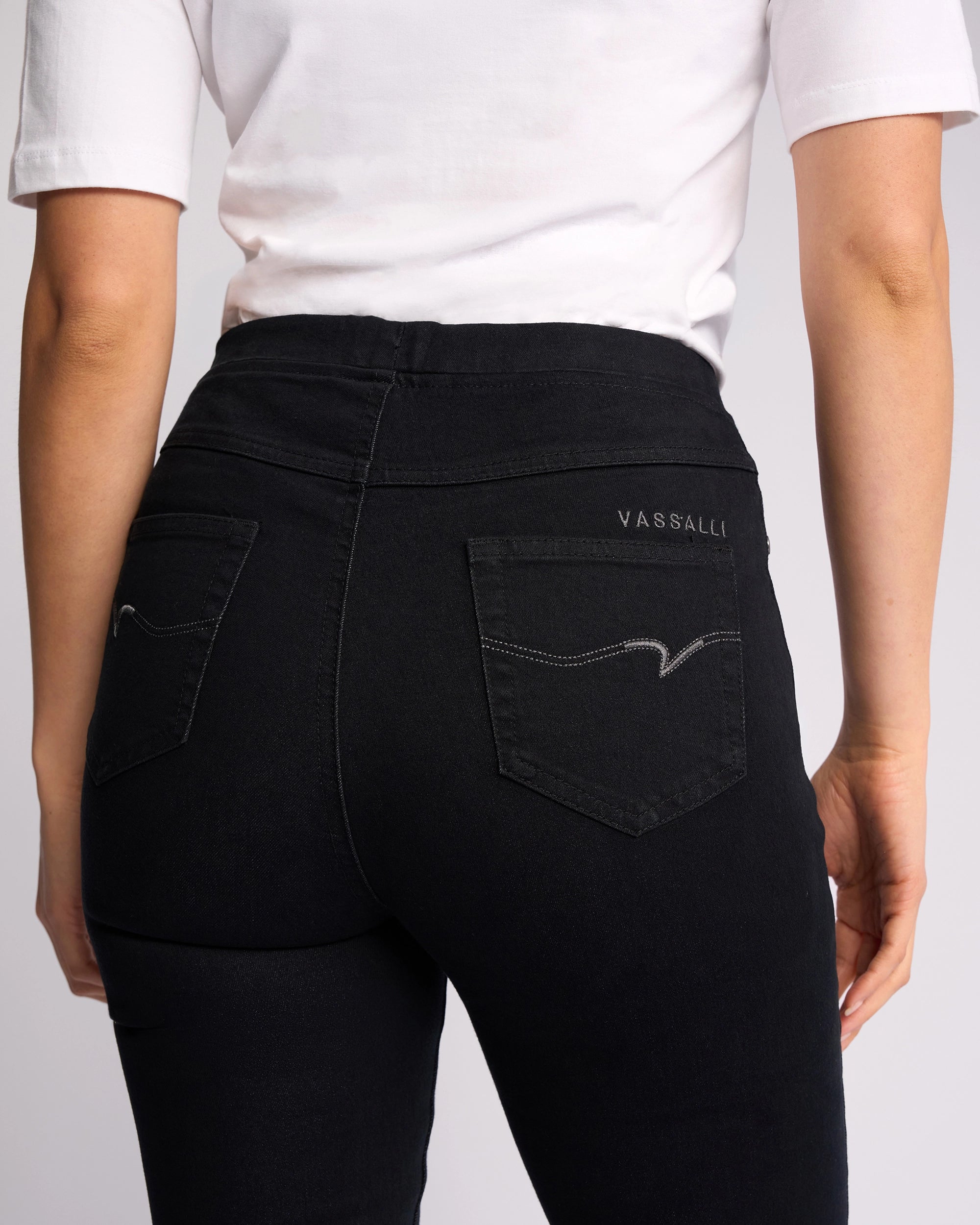 A person seen from the back wearing a white short-sleeve top and Vassalli Bay Pull On Black Denim slim-leg jeans, featuring "Vassalli" embroidered on the right back pocket.