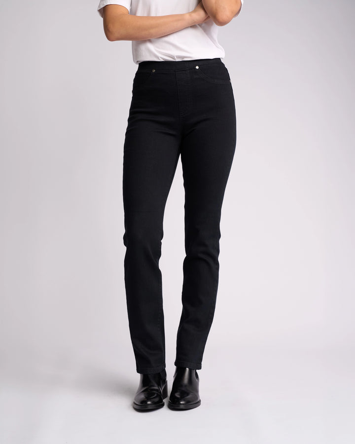 A person stands with arms crossed against a plain light background, wearing a white t-shirt and Vassalli Bay Pull On - Black Denim slim-leg jeans. Face is not visible; black ankle boots complete the look.