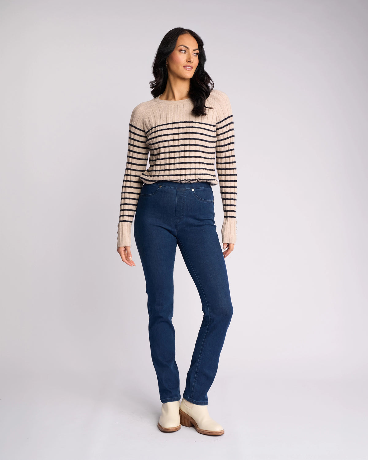A woman with long dark hair wears Vassalli Bay Pull On - Blue Denim (Without Front Fade) jeans, a beige and black striped sweater, and light-colored ankle boots. She stands against a plain light background, looking slightly to the side.