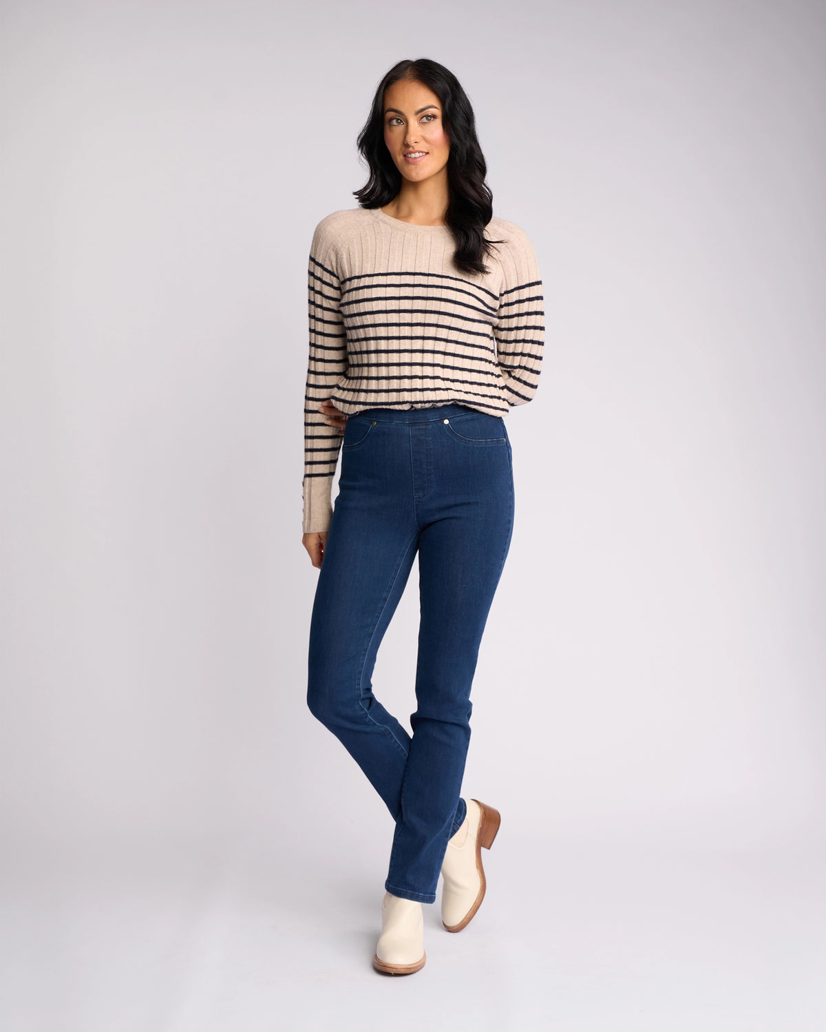 A woman with long dark hair wears the Vassalli Bay Pull On - Blue Denim (Without Front Fade) jeans, a beige sweater with black stripes, and white ankle boots. She stands against a light background, one hand by her side and the other touching her hair.